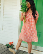 Woman in a red and white checkered dress holding flowers on a wooden deck with a green door.