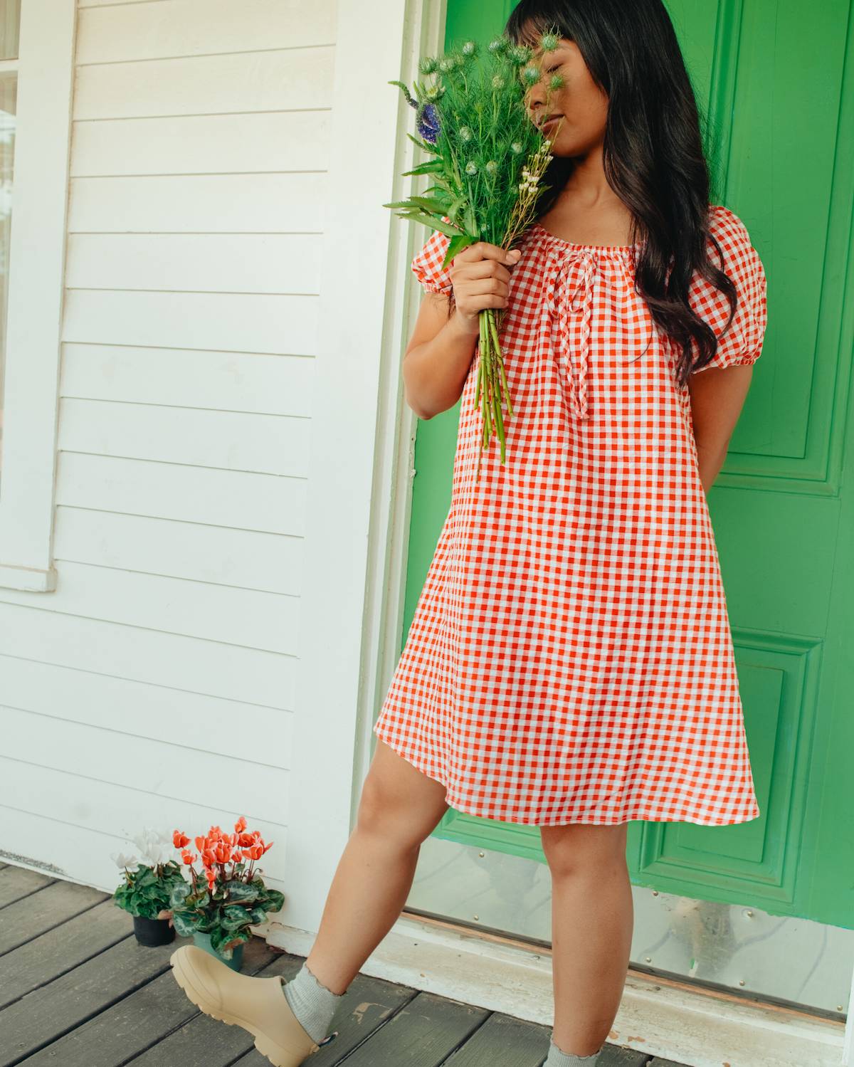 Woman in a red and white checkered dress holding flowers on a wooden deck with a green door.