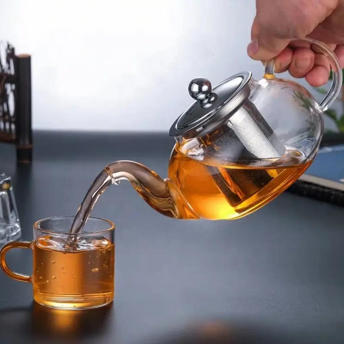 Glass teapot pouring tea into a glass mug on a dark surface.