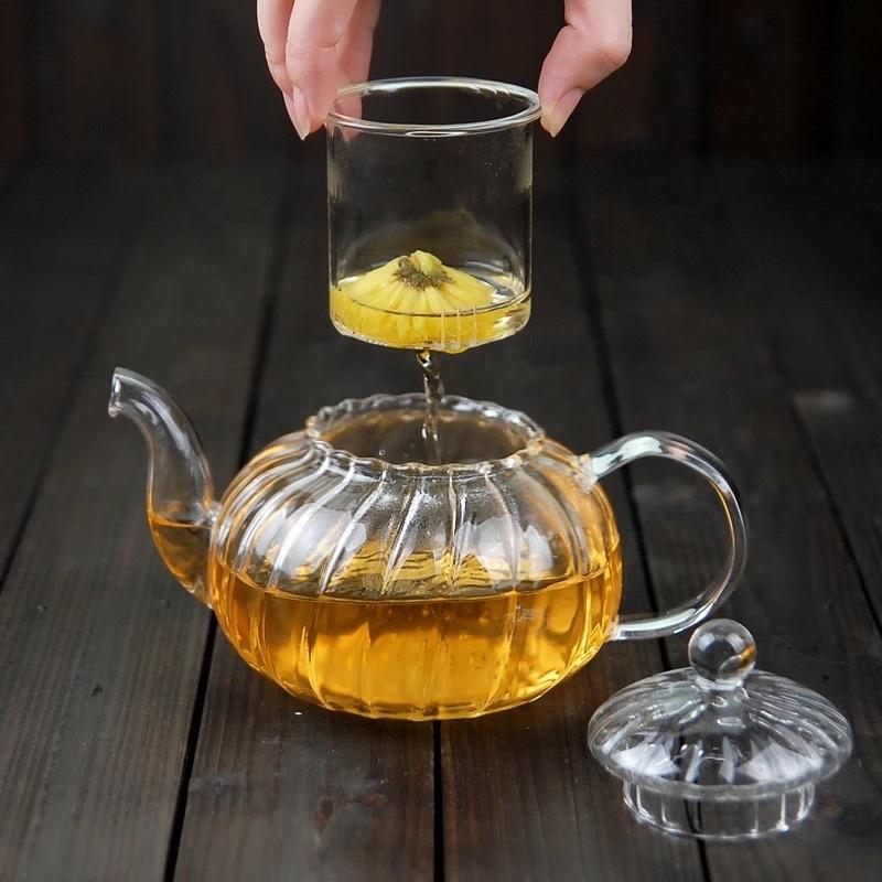 Clear glass teapot with a person pouring tea into a clear glass cup on a wooden surface.