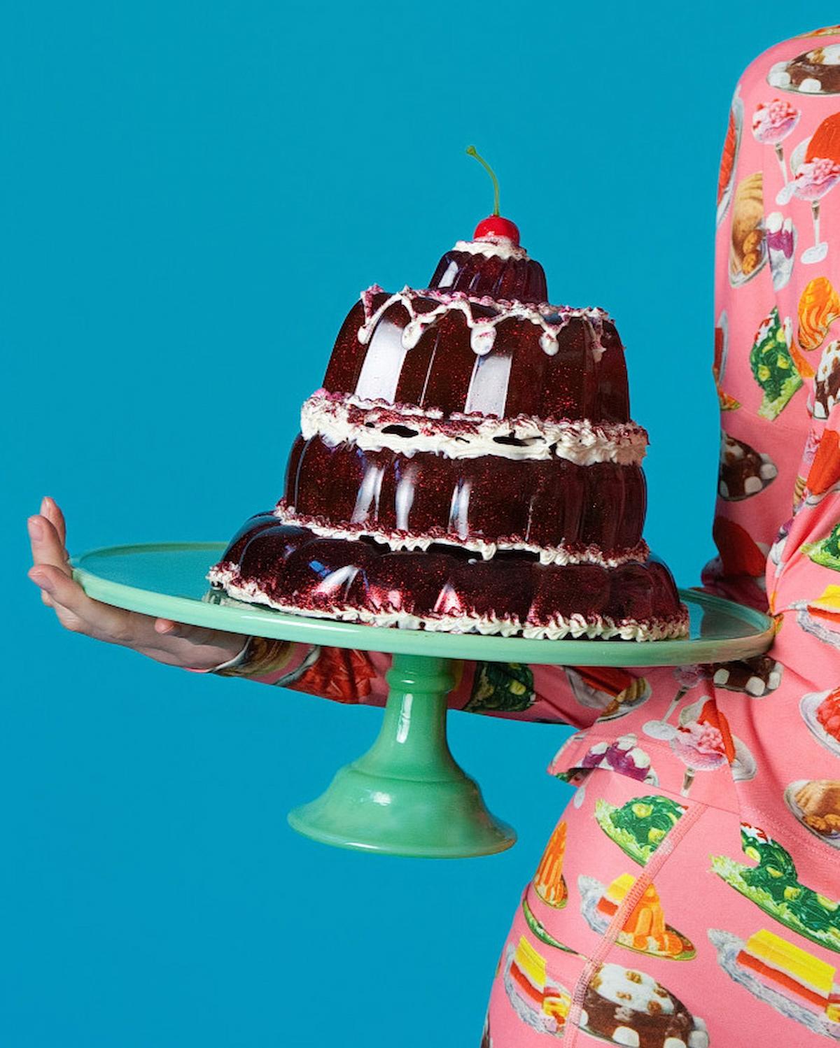 Person holding a three-tiered chocolate cake with white frosting against a blue background