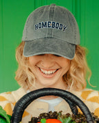 Person wearing a cap with 'HOMEBODY' text, holding a basket of vegetables against a green background