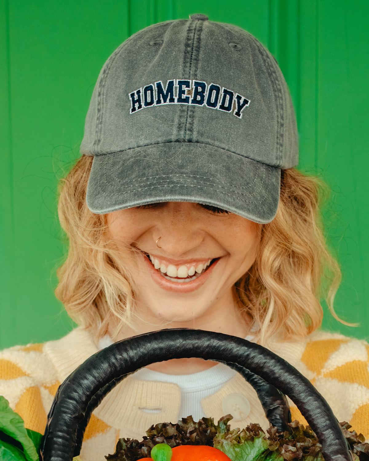 Person wearing a cap with 'HOMEBODY' text, holding a basket of vegetables against a green background
