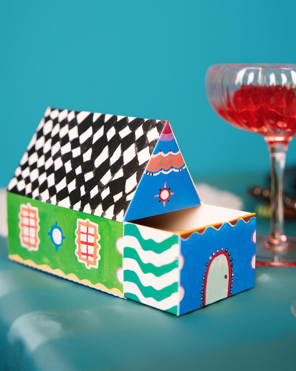 Colorful house-shaped box on a blue surface with a glass of red wine in the background.