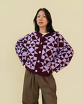 Person modeling a plum and light purple geometric patterned cardigan with white buttons, paired with brown pants, against a solid beige background.