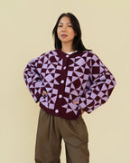 Person modeling a plum and light purple geometric patterned cardigan with white buttons, paired with brown pants, against a solid beige background.