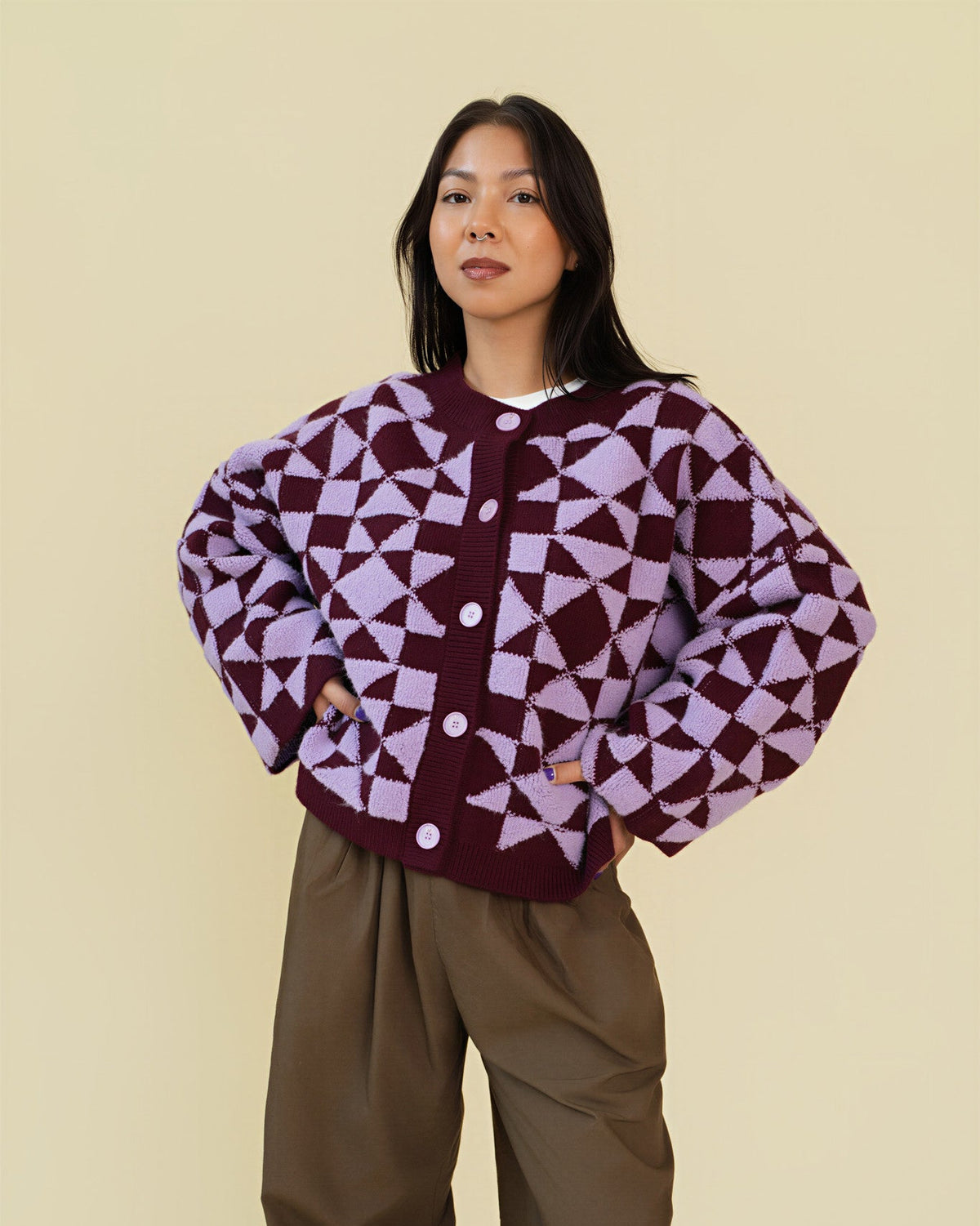 Person modeling a plum and light purple geometric patterned cardigan with white buttons, paired with brown pants, against a solid beige background.