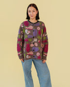 A model wearing a brown button-up cardigan with a colorful vegetable pattern in pink, purple, green, and white, paired with wide-leg blue jeans and black shoes, against a solid pale yellow background.