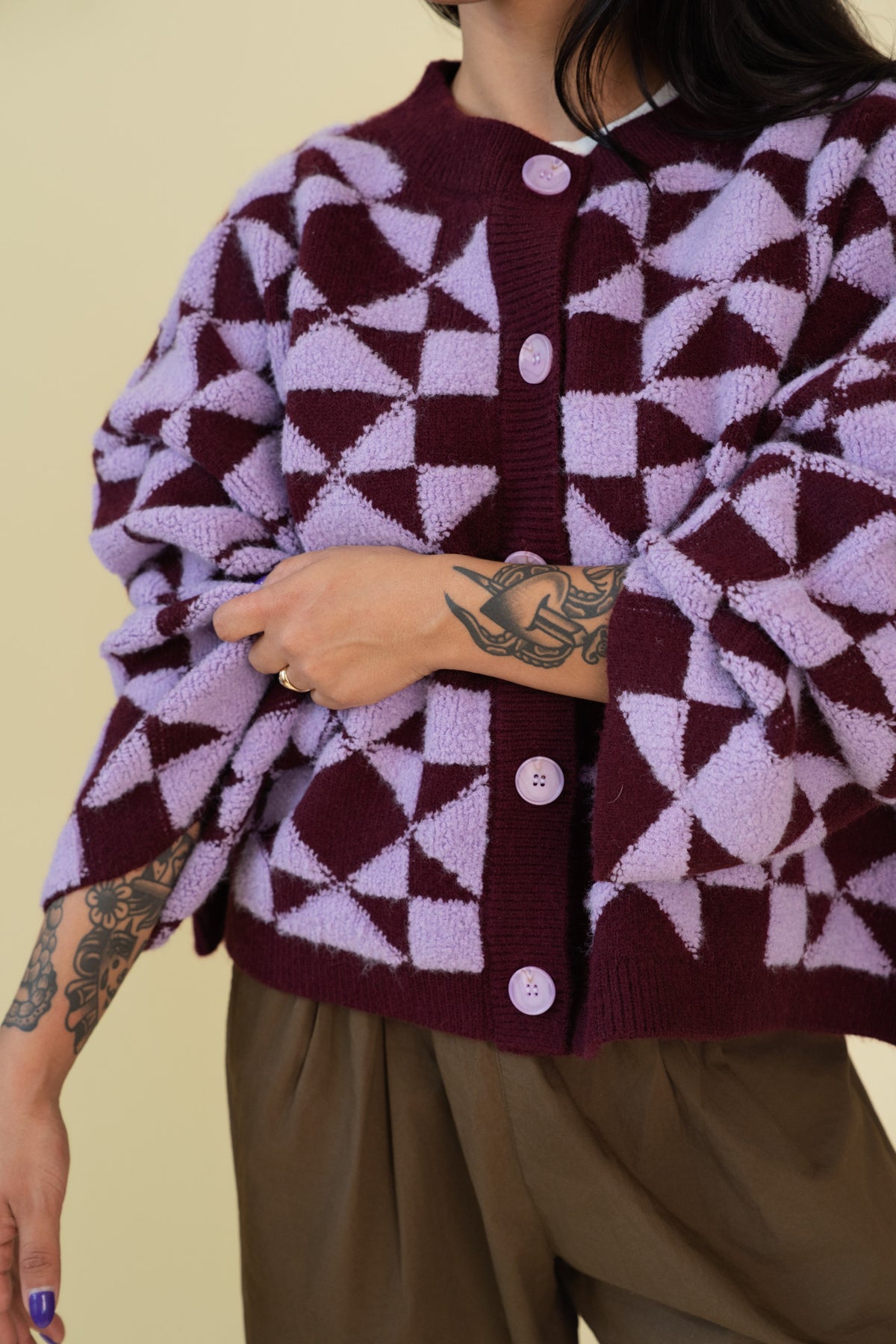 Person modeling a plum and light purple geometric patterned cardigan with white buttons, paired with brown pants, against a solid beige background.