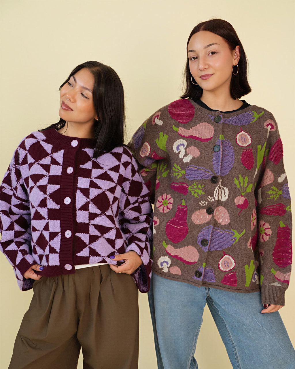 Two women modeling knit cardigans against a plain beige studio background. The woman on the left wears a burgundy and lavender geometric patterned cardigan with olive pants.