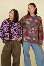 Two people wearing purple knit sweaters and jeans against a pale background. 
