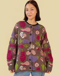 A woman wearing a brown knit cardigan with a colorful vegetable pattern, paired with light blue jeans, against a solid beige background.