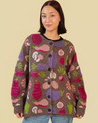 A woman wearing a brown knit cardigan with a colorful vegetable pattern, paired with light blue jeans, against a solid beige background.
