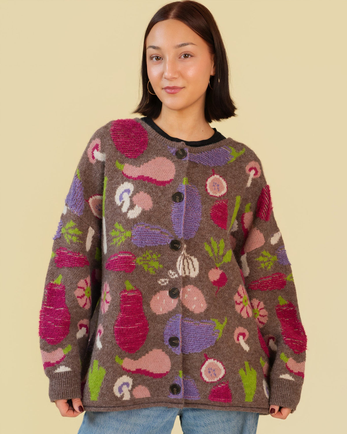 A woman wearing a brown knit cardigan with a colorful vegetable pattern, paired with light blue jeans, against a solid beige background.