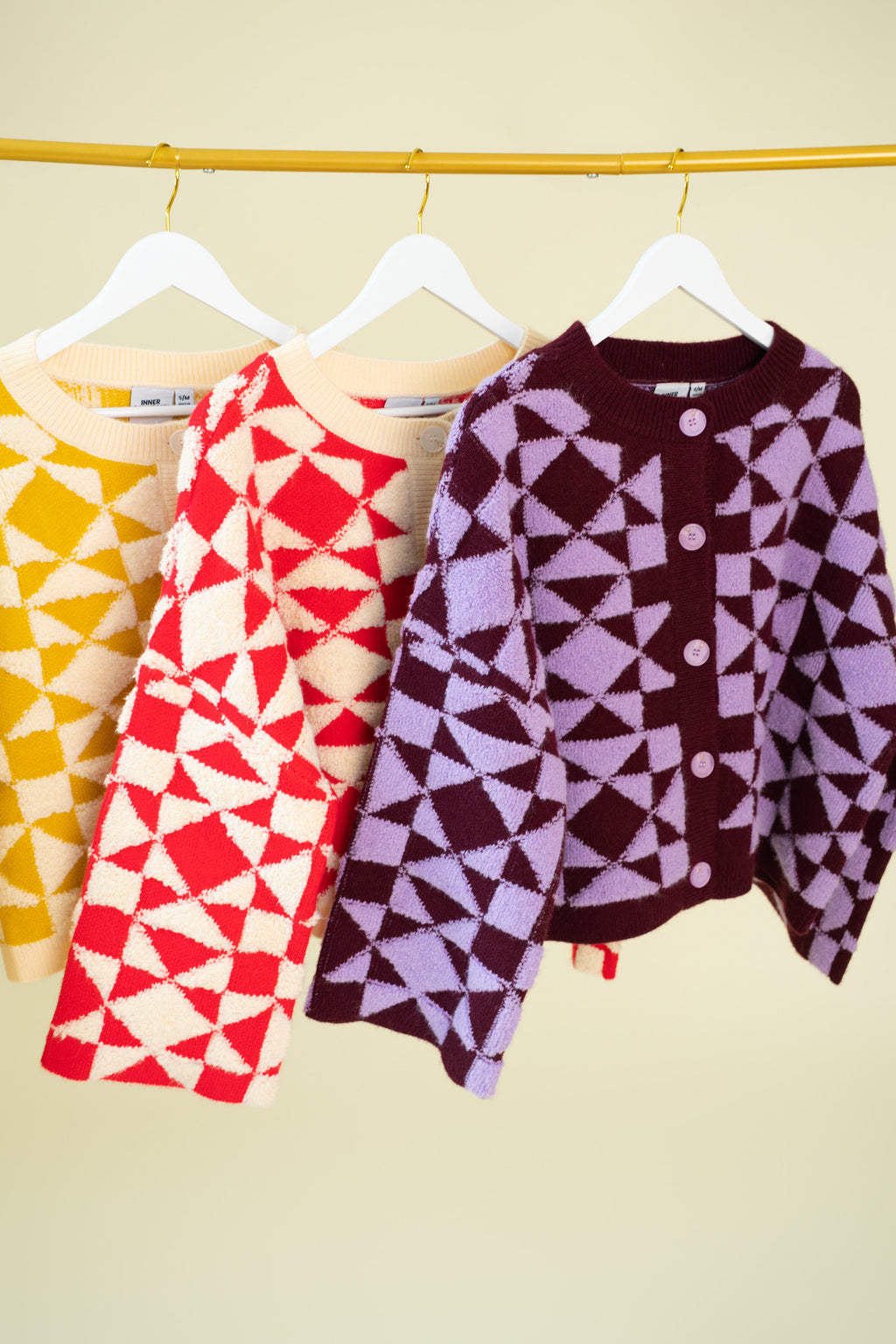 Three geometric patterned sweaters hanging on a gold bar. One sweater is yellow and beige, one is red and cream, and the other is plum and lilac. 