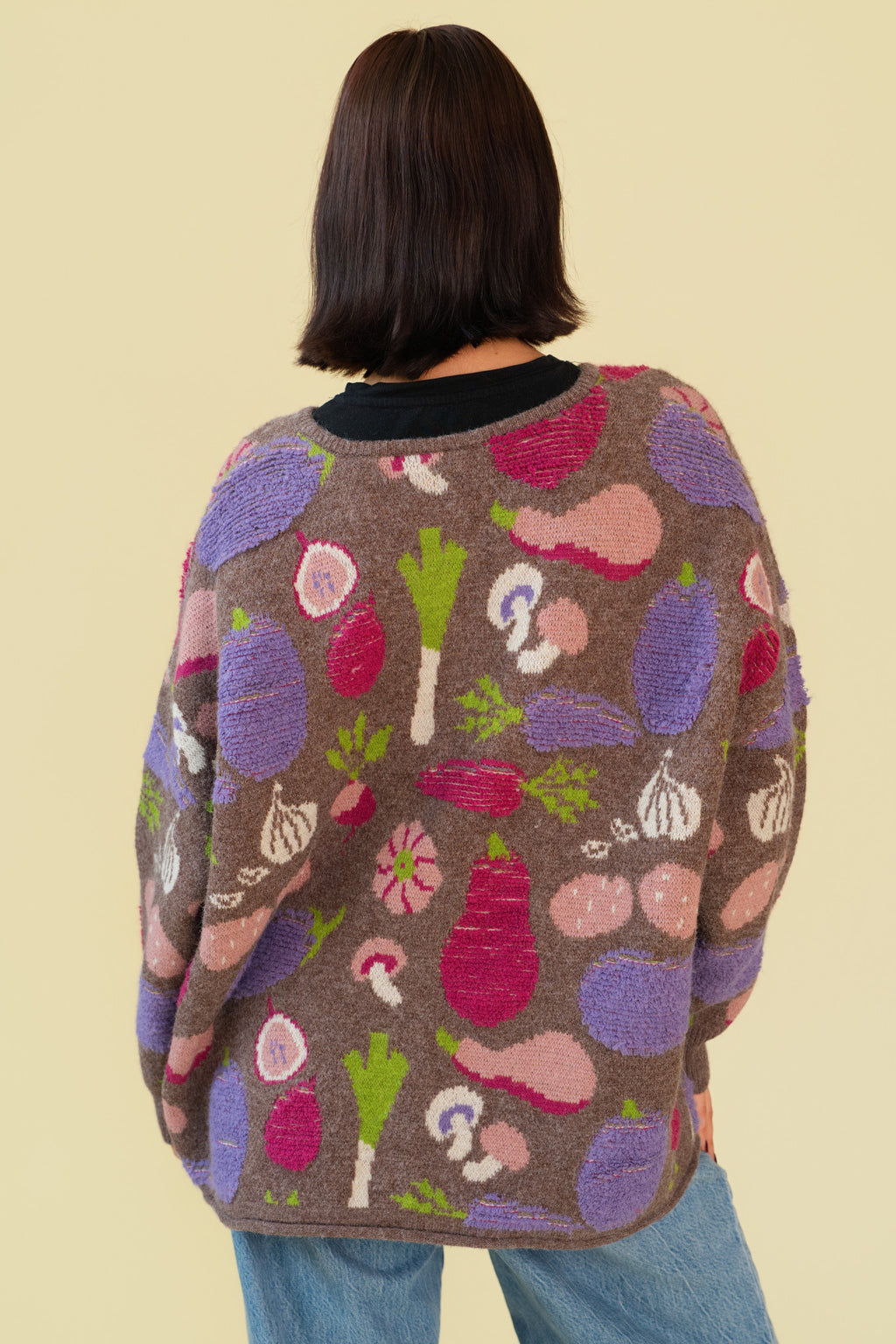 Back of person wearing a chunky knit cardigan sweater with a multicolor vegetable pattern on a brown base, shown against a plain light beige background.