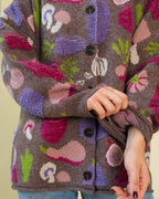 A close-up of a person wearing a chunky knit cardigan sweater with a multicolor vegetable pattern on a brown base. The sweater features black buttons and is shown against a plain light beige background.