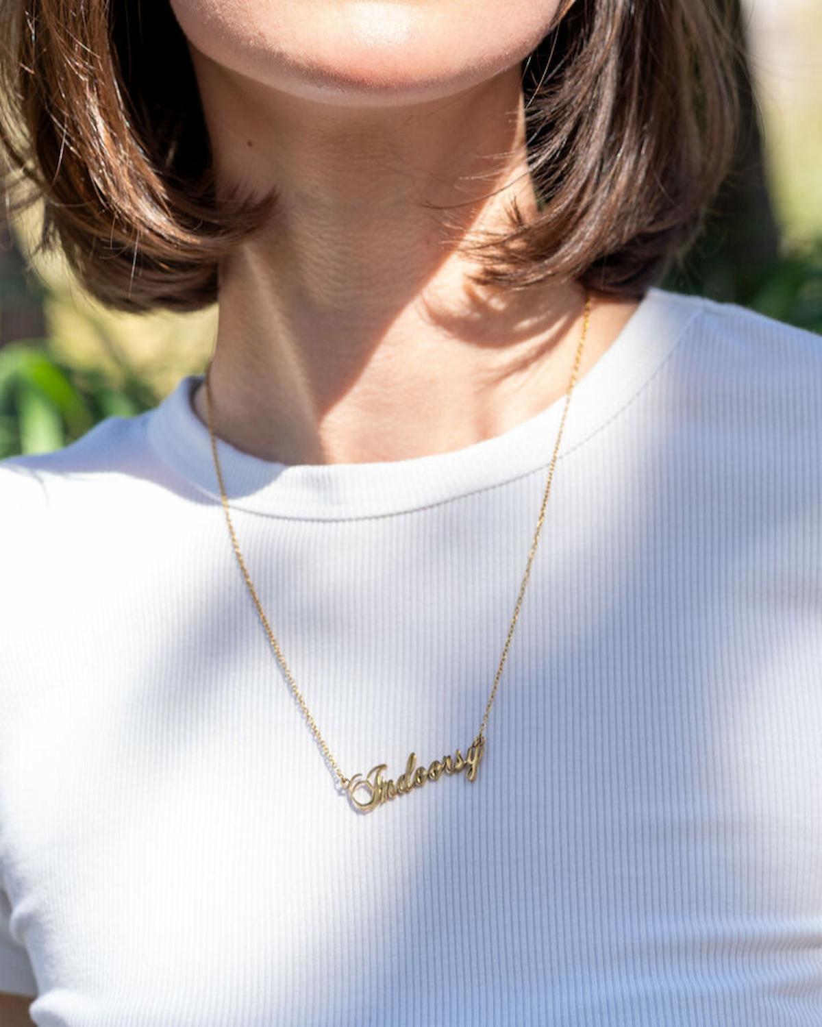 Person wearing a gold necklace with 'Sasha' on a white shirt outdoors