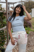 Woman wearing a light blue t-shirt and patterned pants standing in an outdoor garden setting.