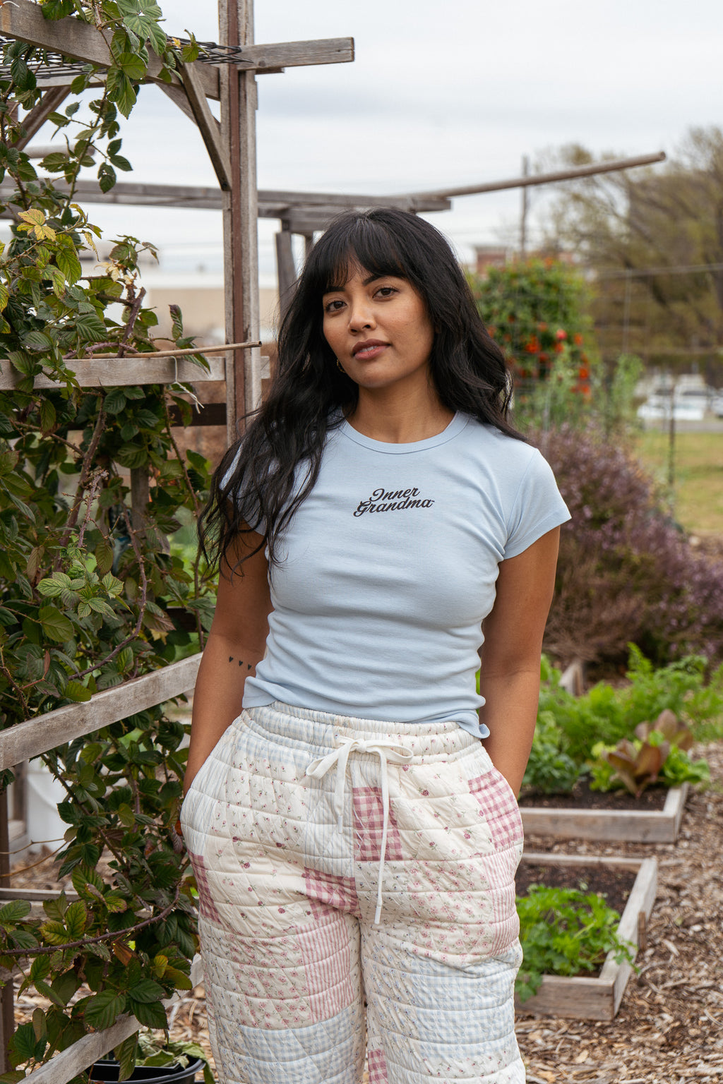 Woman wearing a light blue t-shirt with text and patterned pants standing in a garden.