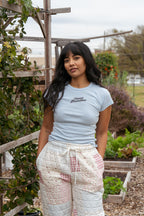Woman wearing a light blue t-shirt with text and patterned pants standing in a garden.