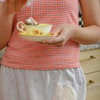 Person wearing a red gingham top and gray pants with lace detail, holding teacup