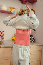 Person wearing a light gray sweatshirt with a doily design and a red checkered shirt underneath, standing in a kitchen.