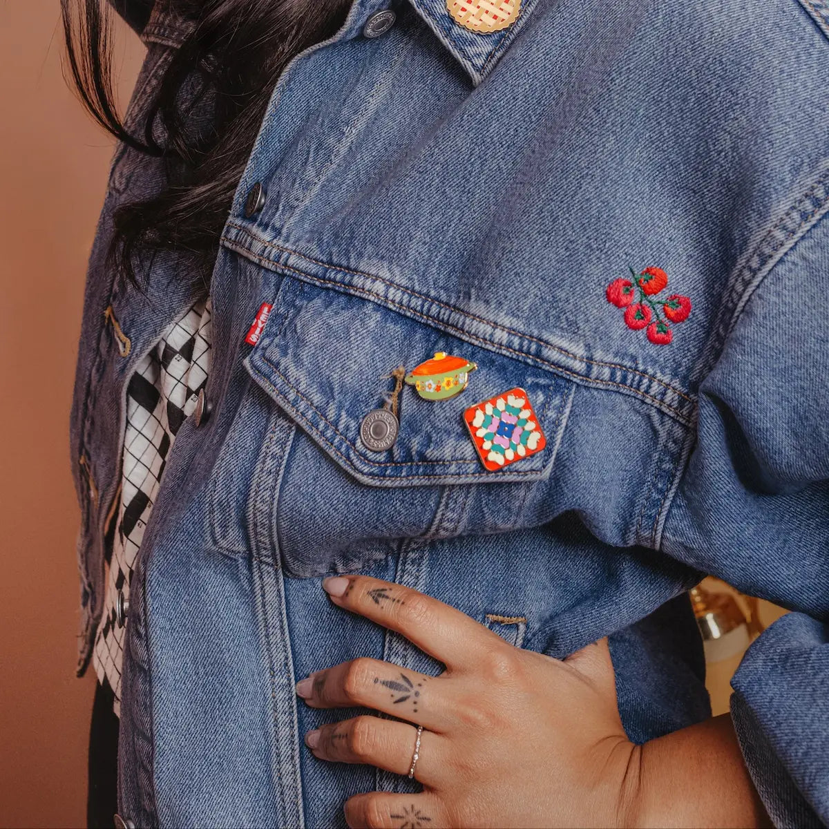 Woman wearing a denim jacket with hot dish pin and grandma quilt pin against a beige background