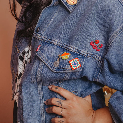Woman wearing a denim jacket with hot dish pin and grandma quilt pin against a beige background