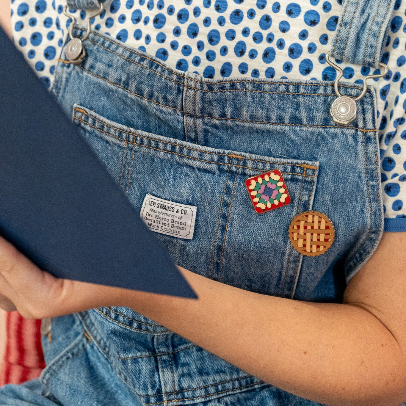 Person wearing a blueberry pattern shirt and denim overalls with pie pin and grandma quilt square pin.