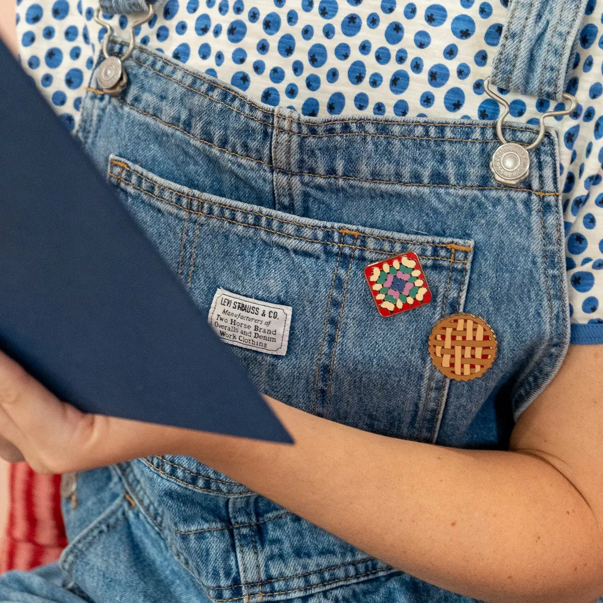 Person wearing blue denim overalls with decorative pins, holding a dark blue folder.