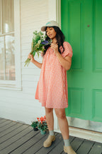 Woman in a red gingham dress holding flowers on a wooden deck with a green door.