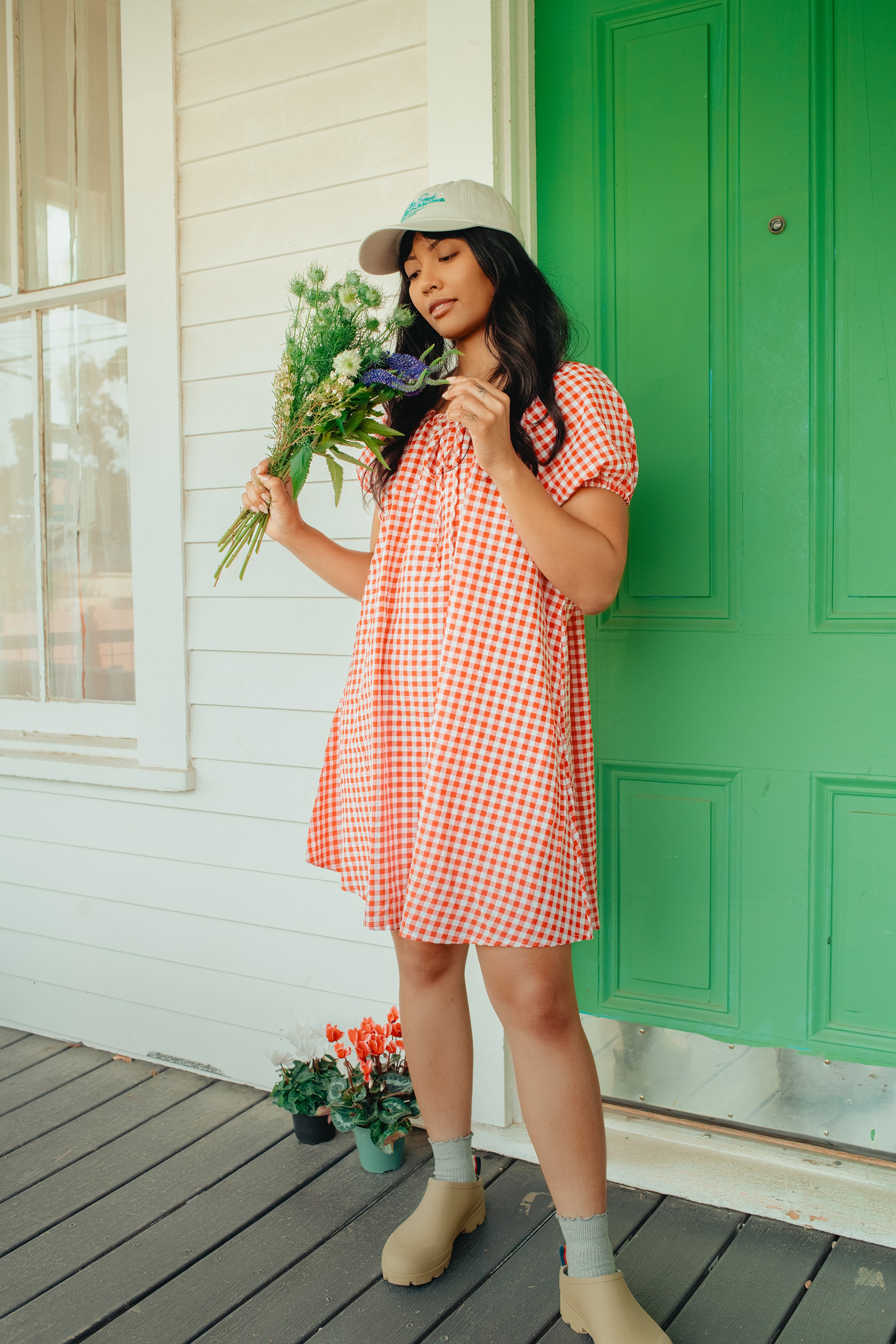 Woman in a red gingham dress holding flowers on a wooden deck with a green door.