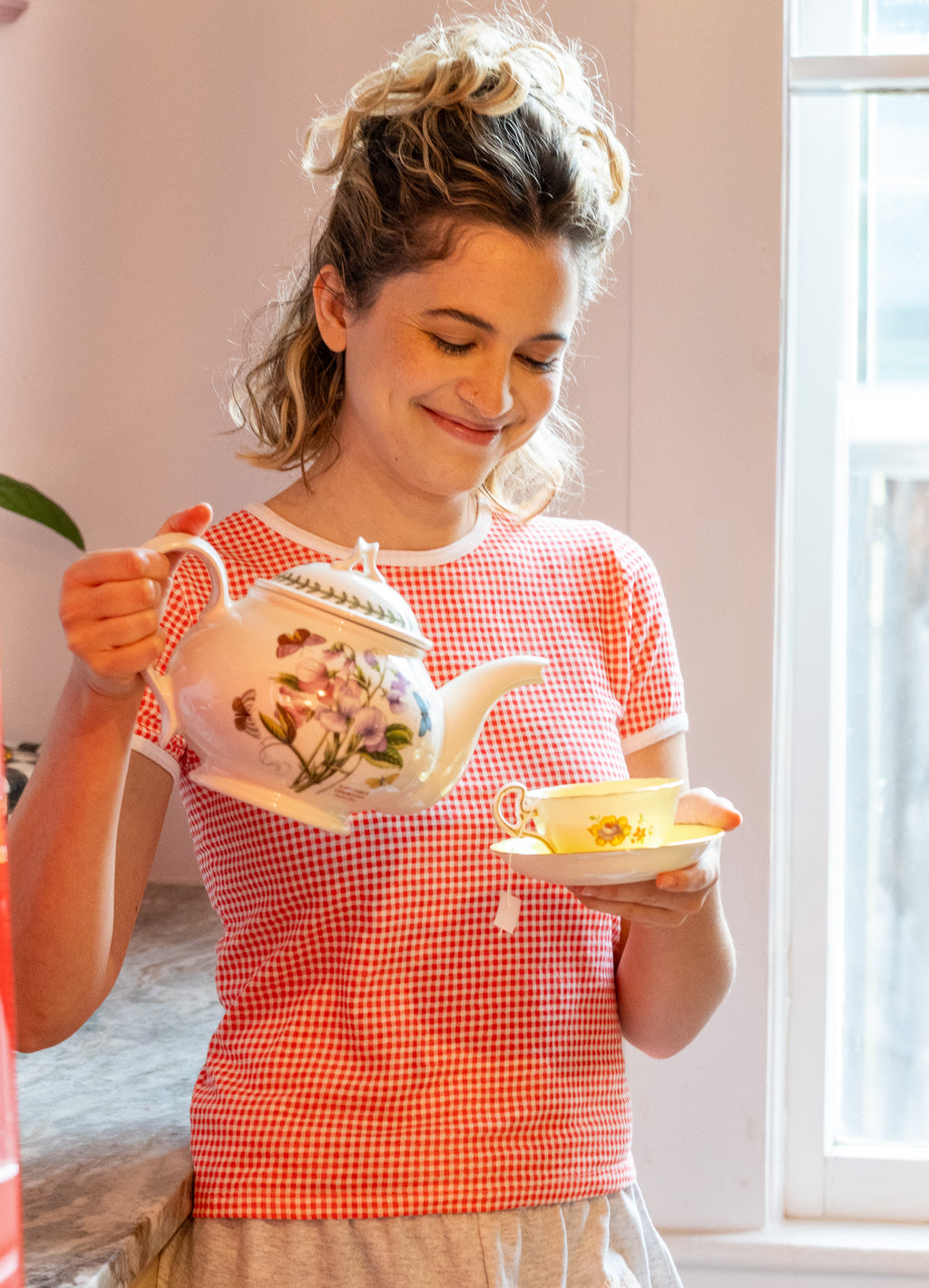 Woman in a red gingham shirt holding a teapot and teacup indoors.
