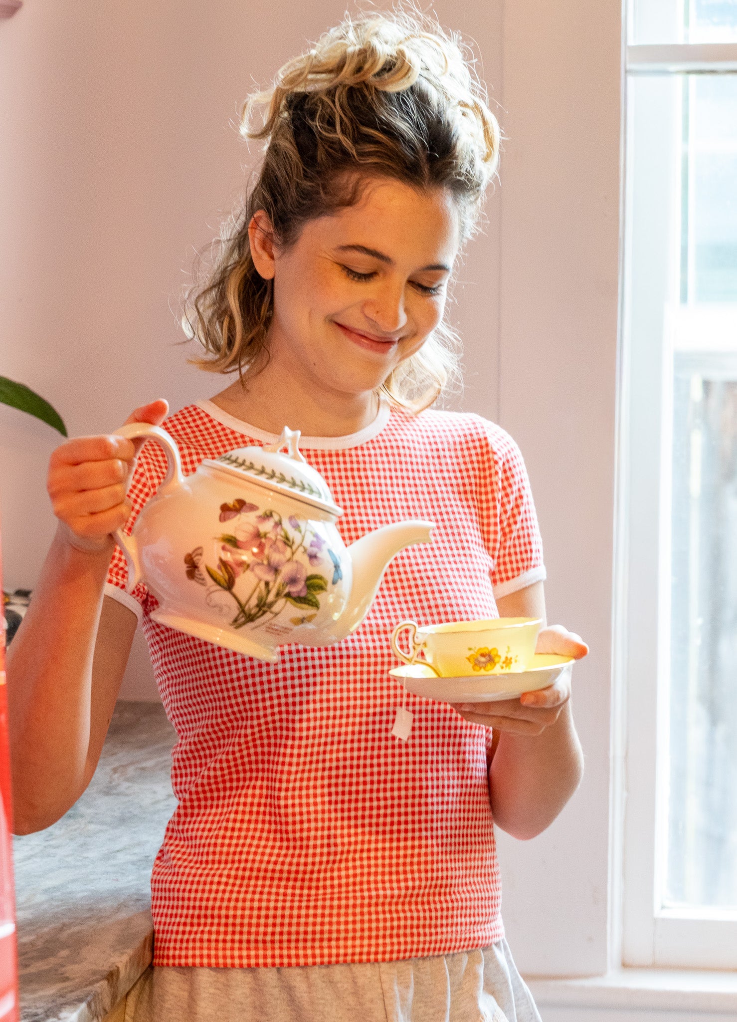Woman in a red gingham shirt holding a teapot and teacup indoors.