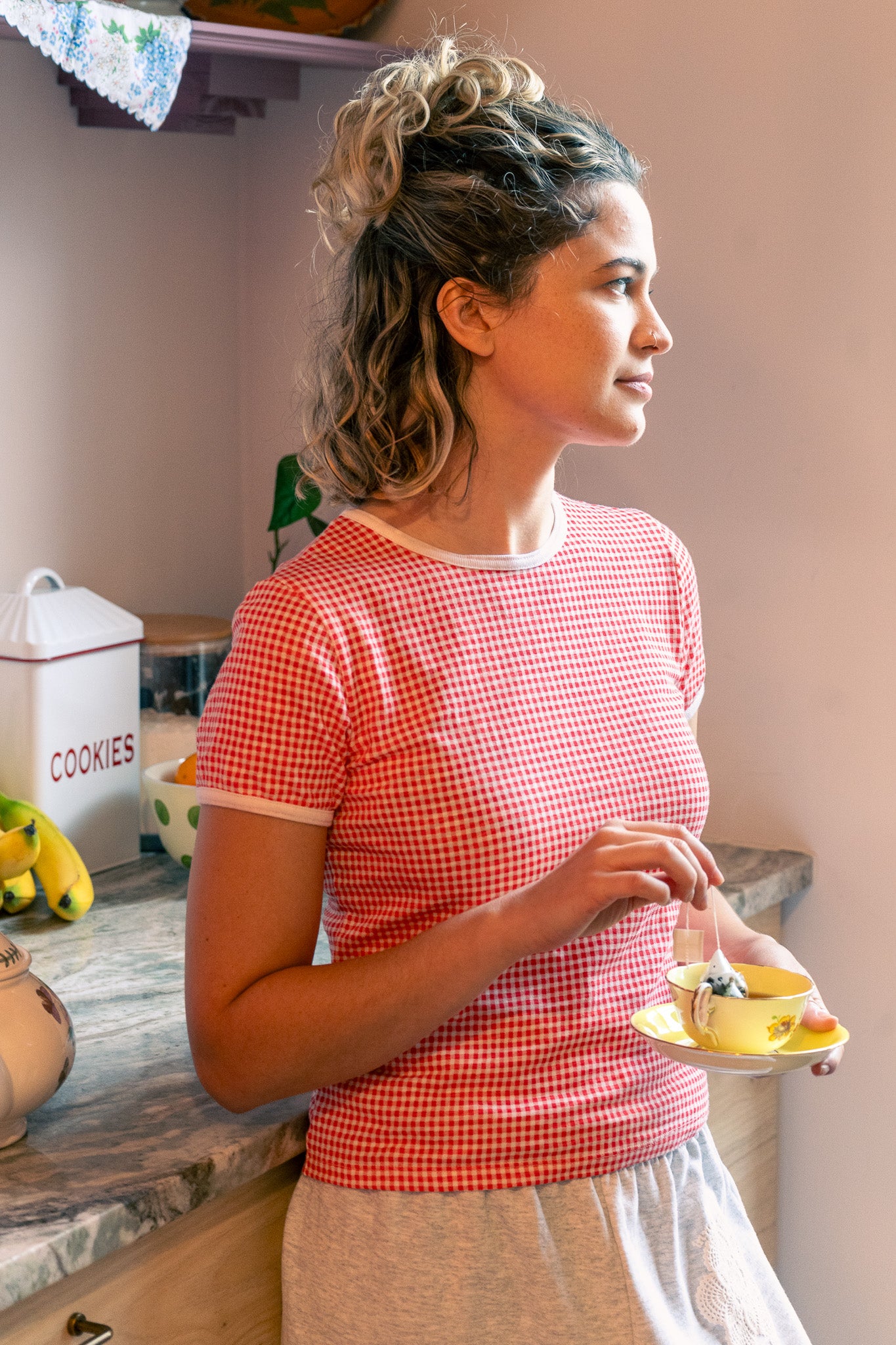 Woman in a kitchen wearing a red gingham tee holding a teacup. 