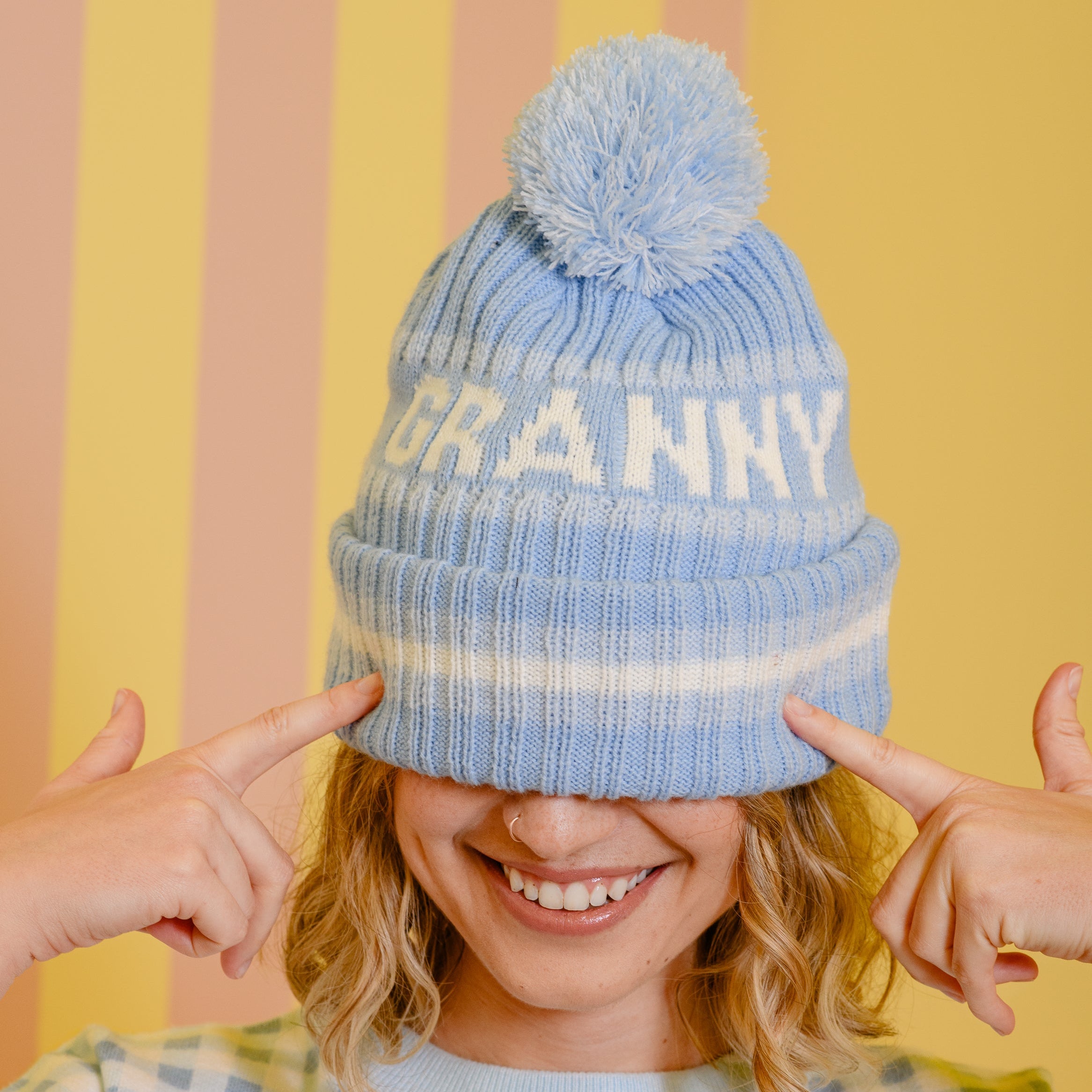 Person wearing a blue 'GRANNY' beanie with a pom-pom against a striped background