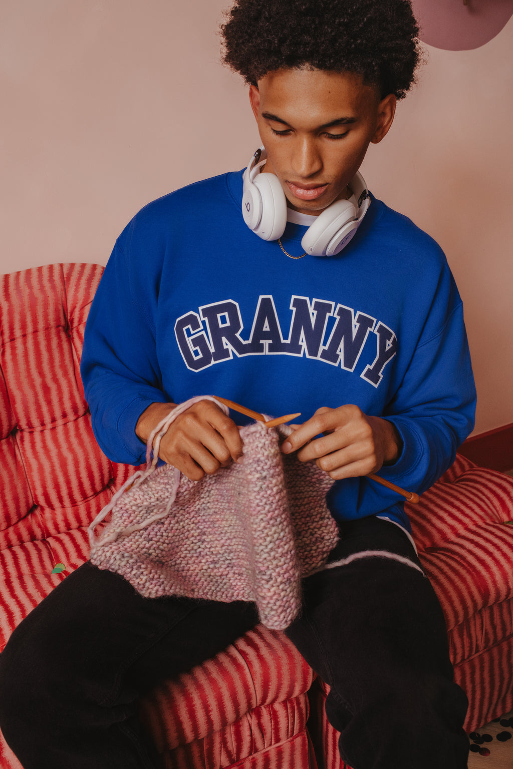 Person wearing a blue sweatshirt with 'GRANNY' text, sitting on a red checkered chair, knitting.