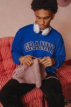 Person wearing a blue sweatshirt with 'GRANNY' text, sitting on a red checkered chair, knitting.