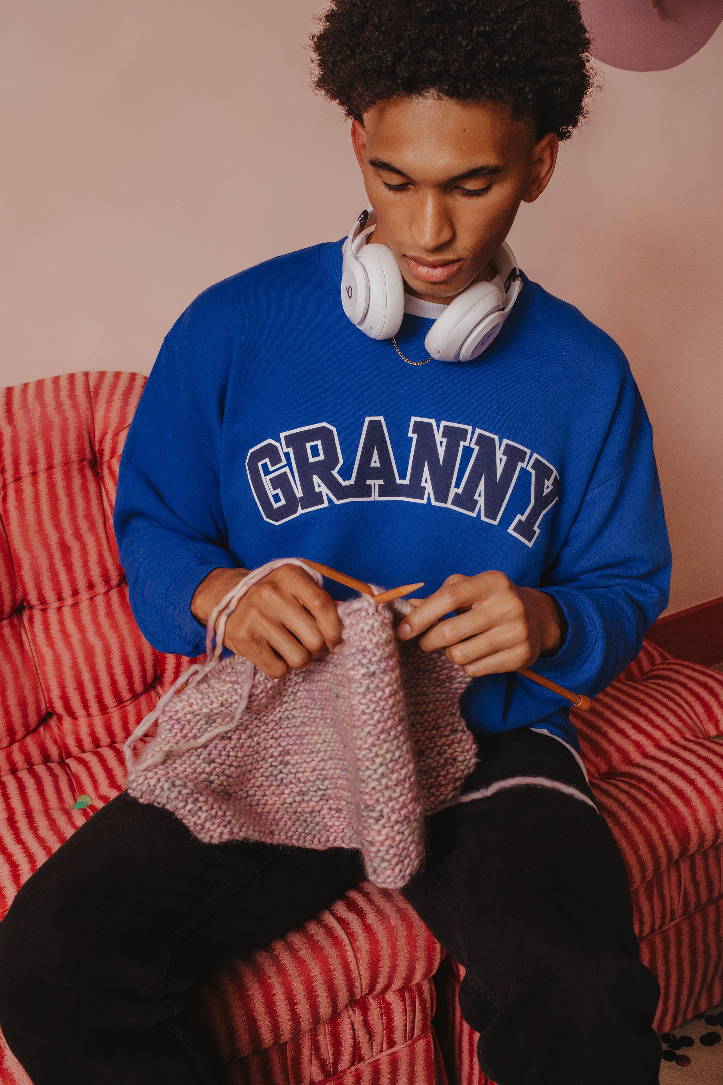 Person wearing a blue sweatshirt with 'GRANNY' text, sitting on a red checkered chair, knitting.