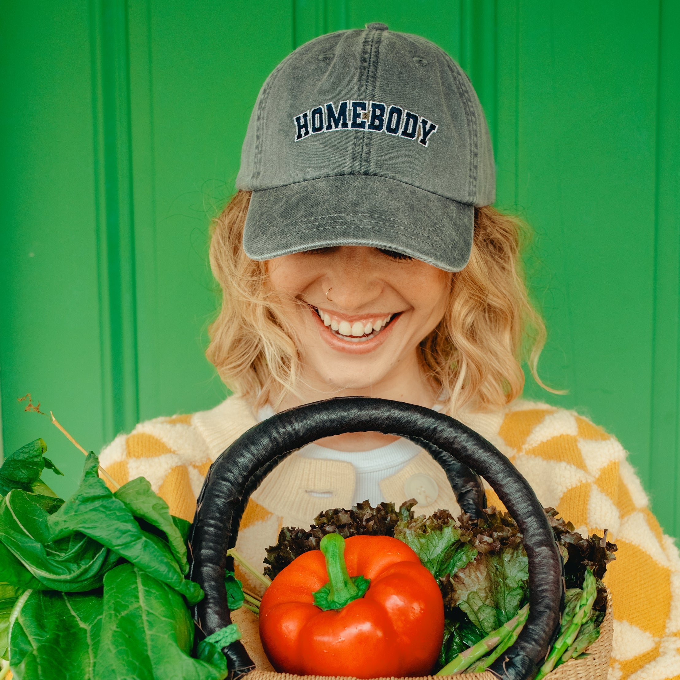 Person wearing a baseball cap with the saying "homebody" holding a woven basket of vegetables against a green background