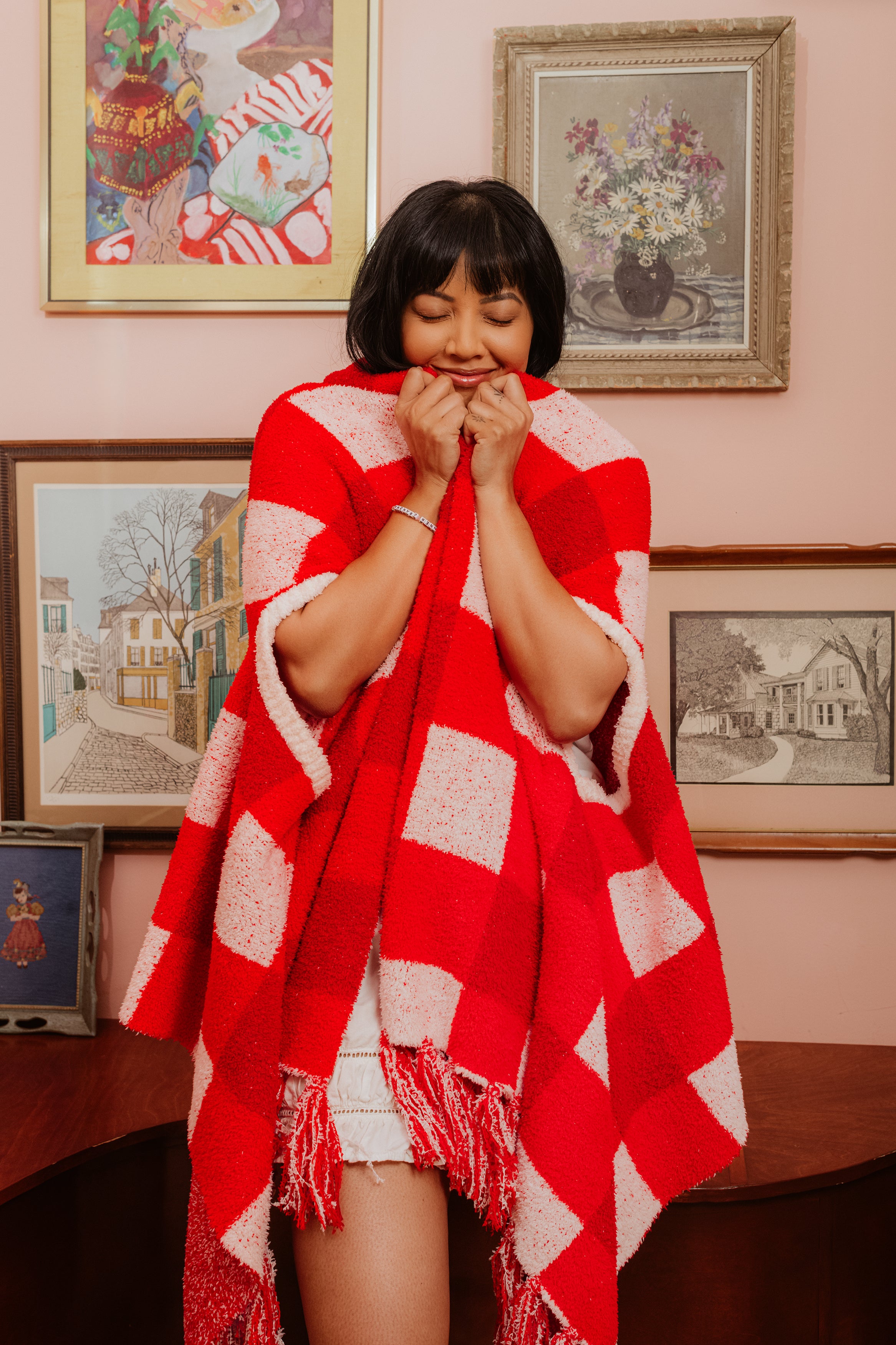 Woman wrapped in a red and white patterned shawl in a room with framed pictures on the wall.