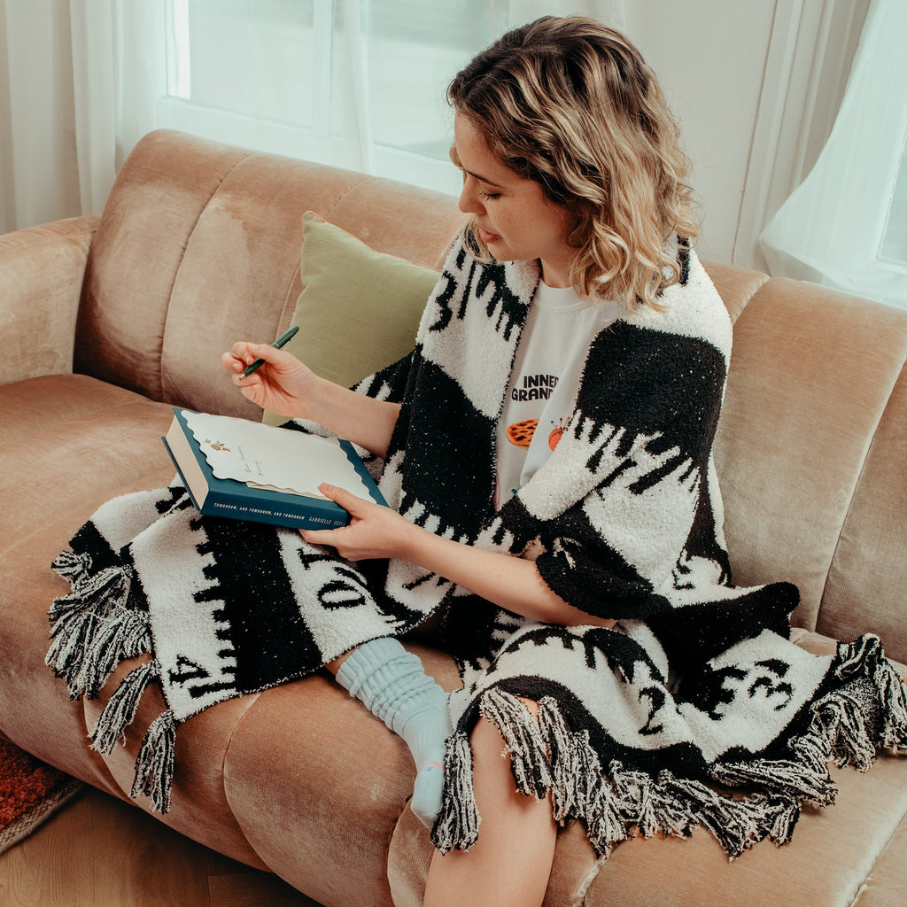 Woman sitting on a couch with a cozy shawl, reading a book in a cozy living room.