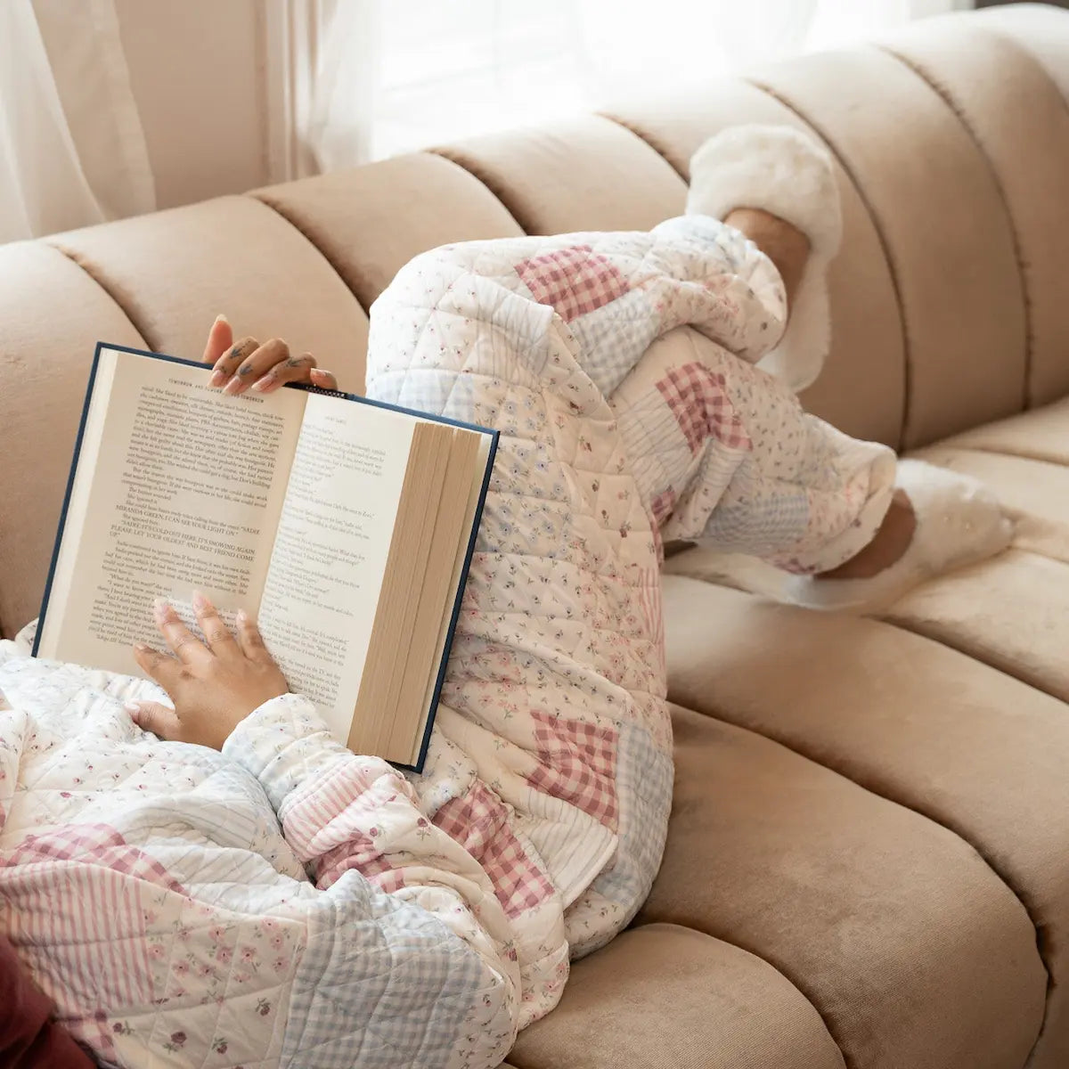 Person reading a book on a beige couch with a book in patchwork sweatpants and white slippers