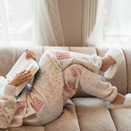Person sitting on a couch with a blanket and book, surrounded by soft light wearing white slippers with pearl details