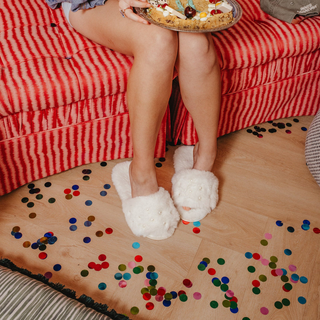 Person sitting on a red striped couch holding a pizza, with confetti on the floor wearing white open toe slippers with pearl details 