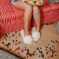 Person sitting on a red striped couch holding a pizza, with confetti on the floor wearing white open toe slippers with pearl details 