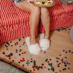 Person sitting on a red striped couch holding a pizza, with confetti on the floor wearing white open toe slippers with pearl details 