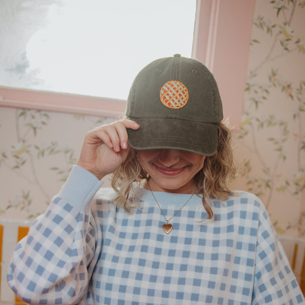 Person wearing a olive green baseball with a pie patch and a blue checkered sweater in a room with floral wallpaper.