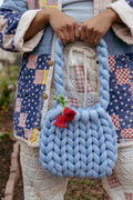 Light Blue large knit bag being held against a womens body with crochet red flower handing from handle